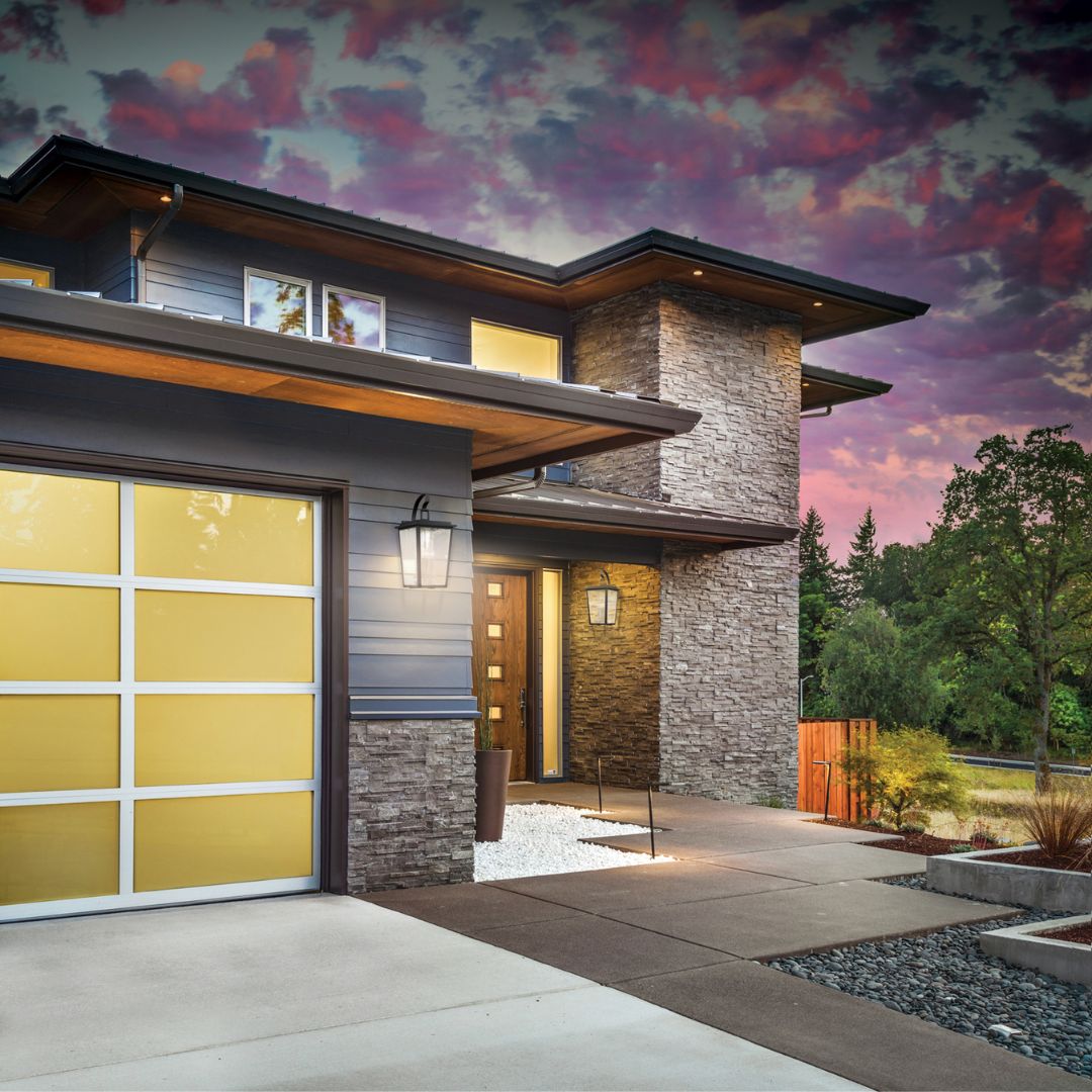 A modern two-story home at sunset with stone and gray siding, featuring large wall-mounted lantern lights by the garage and front door.